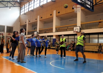 Na zdjęciu przedstawiono moment sportowej konkurencji na hali gimnastycznej. Kilka osób stoi pod koszem do koszykówki i obserwuje lecącą piłkę, która znajduje się tuż obok obręczy.  Wśród uczestników są zarówno zawodnicy w kolorowych koszulkach i szarfach, jak i organizatorzy w żółtych kamizelkach odblaskowych. 