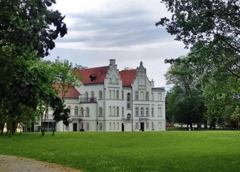 Na zdjęciu widać okazały, jasny pałac lub zabytkowy dwór otoczony rozległym parkiem. Budynek ma białą elewację, czerwony dach oraz ozdobne szczyty i wysokie okna, co nadaje mu elegancki, historyczny charakter.  Przed budynkiem rozciąga się duży zielony trawnik, a całość otaczają drzewa tworzące spokojną, parkową scenerię. Nad pałacem unoszą się ciężkie, szare chmury, które dodają fotografii nastrojowego klimatu.