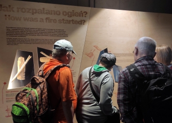 Na zdjęciu widać grupę ludzi stojących przy ścianie ekspozycyjnej. Na ścianie znajduje się tablica z tytułem „Jak rozpalano ogień?” Na ekspozycji widoczne są również ilustracje i prawdopodobnie repliki narzędzi używanych do rozniecania ognia, np. kije, drewienka czy tłoki. Widzowie są skupieni na oglądaniu eksponatów i czytaniu informacji.