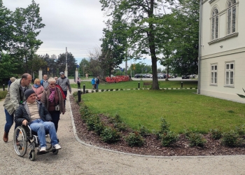 Na zdjęciu widać grupę ludzi spacerujących po zadbanym terenie przy jasnym budynku otoczonym zielenią. Na pierwszym planie mężczyzna pcha wózek inwalidzki, na którym siedzi młody mężczyzna w czapce i jasnej bluzie. Obok nich idzie starsza kobieta w okularach i różowym szaliku. W tle znajdują się kolejne osoby spacerujące alejką, drzewa oraz parking z samochodami. Pogoda jest pochmurna, ale otoczenie wygląda spokojnie i przyjemnie.