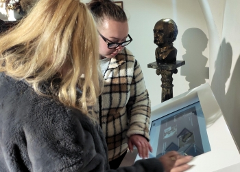 Na zdjęciu widać dwie kobiety korzystające z interaktywnego ekranu na wystawie. Jedna z nich ma blond włosy i ciemne futrzane okrycie, druga nosi okulary oraz jasną kurtkę w kratę. Obie skupiają uwagę na dotykowym panelu prezentującym zdjęcia lub informacje. W tle znajduje się ciemne popiersie ustawione na ozdobnym postumencie, a światło rzuca wyraźne cienie na ścianę. 