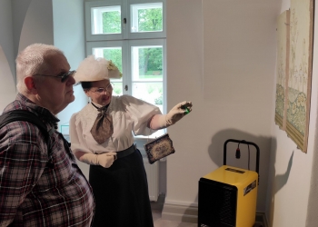 Na zdjęciu widać mężczyznę oraz kobietę ubraną w stylizowany historyczny strój, znajdujących się w sali muzealnej. Kobieta ma biały kapelusz, elegancką bluzkę z bufkami, rękawiczki i małą ozdobną torebkę. Kobieta wskazuje na dekoracyjny fragment sciany. Mężczyzna w okularach przeciwsłonecznych i koszuli w kratę uważnie słucha i obserwuje prezentację. W tle znajduje się duże okno z widokiem na zielony park, a wnętrze jest jasne i spokojne.