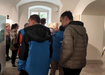 Na zdjęciu widać grupę osób zgromadzonych we wnętrzu muzeum. Ludzie stoją w jasnym pomieszczeniu z łukowymi przejściami i dużym oknem, przez które wpada naturalne światło.W centrum i po prawej stronie stoją mężczyźni w niebieskich i szarych kurtkach, zwróceni w stronę przewodnika lub ekspozycji znajdującej się poza kadrem. Atmosfera wygląda na spokojną i skupioną. 