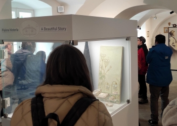 Na zdjęciu przedstawiono wnętrze muzeum z nowoczesnymi gablotami wystawowymi. Na pierwszym planie stoi kobieta w jasnej kurtce i z plecakiem, oglądająca ekspozycję oznaczoną napisem „Piękna historia”. W gablocie znajdują się dekoracyjne elementy i wzorzysta płyta lub kafel. W tle widać grupę zwiedzających spacerujących po sali z łukowymi sklepieniami. Kilka osób zatrzymało się przy innych eksponatach i słucha przewodnika lub ogląda wystawę. Całe wnętrze jest jasne, eleganckie i ma spokojną atmosferę typową dla muzeum.