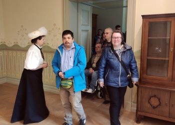 Na zdjęciu widać grupę osób zwiedzających elegancko urządzone wnętrze muzealne. Po lewej stronie stoi kobieta ubrana w historyczny strój z dużym białym kapeluszem. Na środku znajduje się mężczyzna w niebieskiej kurtce, a obok niego kobieta w granatowej kurtce i okularach idzie w stronę aparatu. W tle przez otwarte drzwi widać kolejne osoby, w tym mężczyznę siedzącego na wózku inwalidzkim. Po prawej stronie stoi drewniana, stylowa szafa lub witryna. Pomieszczenie ma jasne ściany z dekoracyjnymi ornamentami i drewnianą podłogę, co nadaje mu zabytkowy i przytulny charakter.