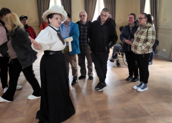 Na pierwszym planie znajduje się przewodniczka ubrana w stylizowany historyczny strój — biały kapelusz, bluzkę z bufkami i długą czarną spódnicę. Kobieta gestykuluje podczas opowiadania, zwracając się do uczestników wycieczki.  Wokół niej stoją zwiedzający w codziennych ubraniach, słuchający z zainteresowaniem. W tle widoczny jest także wózek inwalidzki oraz wysokie okna zasłonięte długimi zasłonami. Sala ma drewnianą podłogę i dekoracyjne ściany, co podkreśla historyczny charakter miejsca.