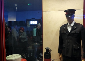 Na zdjęciu widać wystawę poświęconą straży pożarnej, z manekinem ubranym w mundur i hełm strażacki oraz zwiniętą wąż strażacki i sprzęt gaśniczy. W tle przez szybę widać zwiedzających oglądających ekspozycję i interaktywne prezentacje.