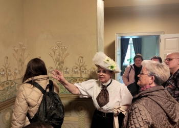 Na zdjęciu widać grupę osób zwiedzających zabytkowe wnętrze muzealne. W centrum znajduje się przewodniczka ubrana w stylizowany historyczny strój z dużym białym kapeluszem ozdobionym kwiatami. Kobieta gestykuluje podczas opowiadania o dekoracyjnej ścianie pokrytej roślinnymi ornamentami.  Wokół niej stoją uczestnicy wycieczki, słuchający z zainteresowaniem. Po lewej stronie widać kobietę z plecakiem odwróconą tyłem do aparatu, a po prawej starsze osoby obserwujące przewodniczkę. W tle znajdują się otwarte drzwi prowadzące do kolejnego pomieszczenia z dużym oknem i zasłonami. Całość tworzy atmosferę spokojnego i edukacyjnego zwiedzania historycznego miejsca.