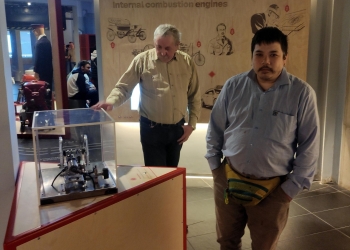 Na zdjęciu widać dwóch mężczyzn znajdujących się w muzeum przy ekspozycji dotyczącej silników spalinowych. Jeden z nich stoi obok gabloty z mechanicznym eksponatem, a drugi patrzy w stronę aparatu; za nimi znajduje się duża plansza edukacyjna. Wnętrze ekspozycji ma nowoczesny charakter, z przyciemnionym oświetleniem i interaktywnymi stanowiskami. W tle można dostrzec innych zwiedzających oraz dodatkowe eksponaty techniczne związane z motoryzacją i historią techniki.