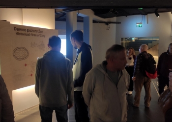 Na zdjęciu widać grupę zwiedzających stojących przed tablicą edukacyjną zatytułowaną „Dawne pożary Żor”. Ekspozycja przedstawia historyczne informacje o pożarach w mieście, a ludzie przyglądają się opisom i ilustracjom, poruszając się po ciemnej sali muzealnej.
