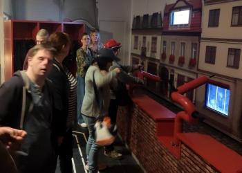 Na zdjęciu grupa osób bierze udział w interaktywnym doświadczeniu w muzeum, używając symulatora strażackiego węża wodnego skierowanego na makietę budynków. Niektórzy uczestnicy mają na głowach czerwone hełmy strażackie, a reszta obserwuje lub czeka na swoją kolej.