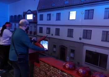 Na zdjęciu widać grupę osób w muzeum, które biorą udział w interaktywnej ekspozycji poświęconej pożarnictwu. Uczestnicy stoją przy symulatorze węża strażackiego, który pozwala „gasić” miniaturową makietę budynków, a część z nich ma na głowach czerwone hełmy strażackie ustawione na barierce. W tle widać detale fasad budynków, w tym okna i dachy, a oświetlenie ekspozycji nadaje scenie realistyczny, niebieskawy efekt, imitujący noc lub sytuację awaryjną. Osoby skupione są na celowaniu strumieniem wody w symulowany pożar, a reszta obserwuje lub przygotowuje się do swojej kolejki.