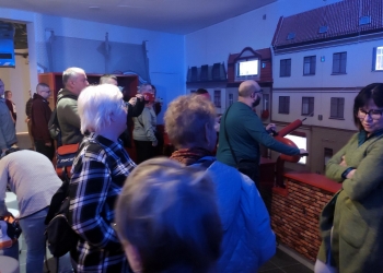 Na zdjęciu widać grupę ludzi w muzeum uczestniczących w interaktywnej ekspozycji pożarniczej, w której można sterować wężem strażackim skierowanym na makietę budynków. W tle widać makiety kamienic z oknami, a niebieskie oświetlenie nadaje scenie wrażenie nocnej akcji ratunkowej, podczas gdy część osób obserwuje, a część aktywnie uczestniczy w symulacji gaszenia pożaru.
