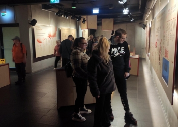 Na zdjęciu widzimy grupę osób w muzeum, stojących przy interaktywnej ekspozycji edukacyjnej o spalaniu i pożarach. Na ścianach znajdują się panele informacyjne z tekstem i ilustracjami, a zwiedzający czytają i oglądają eksponaty, tworząc atmosferę naukowego poznawania historii i mechanizmów pożarów.