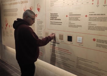Na zdjęciu widać młodego mężczyznę stojącego przy ścianie edukacyjnej w muzeum, na której przedstawiono informacje o rekordowych temperaturach osiąganych przez człowieka oraz historii pomiaru ciepła. Zwiedzający wydaje się badać interaktywny panel z różnymi materiałami, testując, który najlepiej chroni przed ciepłem, a plansza zawiera teksty i ilustracje naukowe w języku polskim i angielskim.