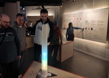 Na zdjęciu widać grupę zwiedzających w muzeum nauki, którzy oglądają interaktywną ekspozycję poświęconą pomiarom temperatury i przewodnictwu ciepła. W centrum kadru znajduje się stojąca lampa w formie cylindrycznego, świecącego słupa zmieniającego kolory, którą najprawdopodobniej można dotykać lub regulować, aby obserwować efekty na otaczających panelach edukacyjnych.