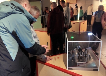 Kilka osób ogląda ekspozycje i korzysta z urządzeń edukacyjnych. Na pierwszym planie dwie osoby stoją przy stanowisku z eksponatem umieszczonym w szklanej gablocie — wygląda on jak mechaniczne lub techniczne urządzenie z metalowymi kołami. Jedna z osób wskazuje palcem panel informacyjny, a druga trzyma telefon.  W tle znajdują się inni zwiedzający, rozproszeni po sali i oglądający kolejne stanowiska.