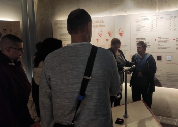 Na zdjęciu przedstawiono grupę osób zwiedzających muzeum. Zwiedzający stoją przy interaktywnej ekspozycji dotyczącej temperatury i skali Celsjusza — na ścianie widać plansze edukacyjne z ilustracjami naukowców, tabelami oraz opisami w języku polskim i angielskim.  Na pierwszym planie znajduje się mężczyzna w szarej bluzie z przewieszoną przez ramię torbą, stojący tyłem do aparatu. Obok niego są inne osoby słuchające lub obserwujące prezentację. Po prawej stronie widać młodego mężczyznę stojącego przy stanowisku interaktywnym z metalowym urządzeniem przypominającym element eksperymentu naukowego.