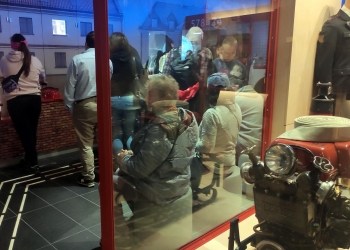 Zwiedzający znajdują się za przeszkloną ścianą, oglądając ekspozycję przypominającą wnętrze pojazdu lub stanowisko edukacyjne, a po prawej stronie widoczny jest zabytkowy sprzęt strażacki z reflektorem i zwiniętym wężem.  W tle można zauważyć elementy scenografii imitujące ulicę lub miejską zabudowę, co nadaje wystawie realistyczny charakter. Całość ma nowoczesną formę ekspozycji interaktywnej, z nastrojowym oświetleniem i licznymi eksponatami technicznymi.