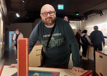 Na zdjęciu widać uśmiechniętego mężczyznę stojącego przy interaktywnym stanowisku w muzeum. Mężczyzna ma okulary, brodę i jest ubrany w ciemnozieloną bluzę z dużym napisem na przodzie oraz przewieszoną przez ramię torbę. Opiera ręce o blat ekspozycji i patrzy prosto w aparat, sprawiając wrażenie zadowolonego i zainteresowanego miejscem.  W tle znajdują się inni zwiedzający korzystający z różnych stanowisk edukacyjnych. Wnętrze ma nowoczesny charakter — ciemny sufit z punktowym oświetleniem kontrastuje z jasnymi ścianami i drewnianymi elementami ekspozycji.