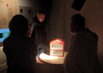 Na zdjęciu widać kilka osób zgromadzonych wokół interaktywnej ekspozycji w muzeum. W centrum znajduje się podświetlony model małego domu lub przekroju budynku ustawiony na okrągłym stanowisku. Jasne światło skierowane na eksponat tworzy kontrast z ciemniejszym otoczeniem sali.  Wokół stanowiska stoją zwiedzający — część z nich obserwuje model, a jedna osoba pochyla się, przyglądając się bliżej ekspozycji. Po lewej stronie widoczna jest ściana z planszami edukacyjnymi zawierającymi tekst i ilustracje dotyczące nauki lub technologii.