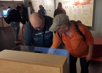Na zdjęciu widać dwóch mężczyzn pochylających się nad interaktywnym eksponatem w muzeum. Jeden z nich ma okulary i ciemną bluzę z nadrukiem, drugi ubrany jest w pomarańczową koszulkę polo oraz jasną czapkę z daszkiem. Obaj z zainteresowaniem zaglądają do wnętrza urządzenia znajdującego się w drewnianej obudowie ze szklaną osłoną.  W tle znajdują się inni zwiedzający oraz duże plansze edukacyjne z tekstami i grafikami dotyczącymi nauki i technologii.