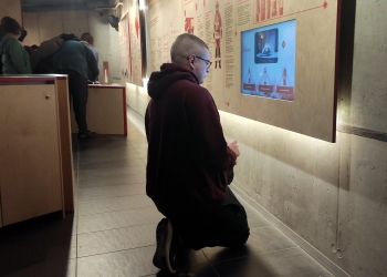 Na zdjęciu widać osobę klęczącą przed interaktywną ekspozycją w muzeum i skupioną na ekranie informacyjnym zamontowanym w ścianie. W tle inni zwiedzający korzystają z wystawy, a nowoczesne, przyciemnione wnętrze podkreśla edukacyjny charakter miejsca.