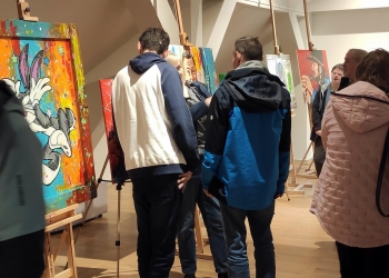 Na zdjęciu widać grupę osób oglądających obrazy podczas wystawy w galerii sztuki. Na pierwszym planie znajdują się kolorowe obrazy inspirowane postaciami z kreskówek i popkultury, ustawione na sztalugach. Uczestnicy wydarzenia rozmawiają i przyglądają się pracom w jasnym, nowoczesnym wnętrzu.