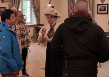 Na zdjęciu widać grupę osób stojących w eleganckim wnętrzu. W centrum znajduje się kobieta ubrana w stylizowany, historyczny strój — biały kapelusz ozdobiony kwiatami, jasną bluzkę i ciemną kokardę pod szyją. Pozostałe osoby są ubrane współcześnie i słuchają jej z uwagą. Pomieszczenie jest jasne, z dużymi oknami, zasłonami i dekoracjami na ścianach, co nadaje miejscu elegancki, spokojny klimat.