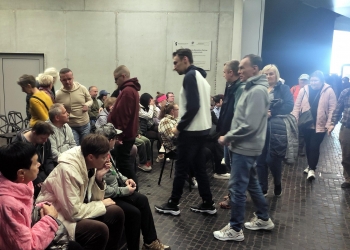 Na zdjęciu widać grupę ludzi zgromadzonych w dużym, nowoczesnym wnętrzu. Część osób siedzi na krzesłach ustawionych w rzędach, inni przechodzą między nimi lub stoją i rozmawiają. Większość ma na sobie kurtki i bluzy. Pomieszczenie ma surowy, industrialny charakter — dominują betonowe ściany i ciemna podłoga z drobnej kostki. Po prawej stronie wpada jasne światło z wejścia lub dużego okna.