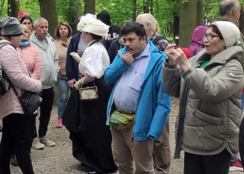 Na zdjęciu widać grupę ludzi zgromadzonych podczas spaceru lub wydarzenia plenerowego w parku pełnym wysokich drzew. Uczestnicy są ubrani w kurtki i cieplejsze ubrania, co sugeruje chłodniejszy dzień, a większość z nich stoi i słucha lub obserwuje coś poza kadrem.  W centrum znajduje się osoba ubrana w stylizowany historyczny strój z dużym jasnym kapeluszem, co nadaje scenie charakter rekonstrukcji historycznej lub tematycznej wycieczki. Obok stoją inni uczestnicy, rozmawiający i przyglądający się wydarzeniu w spokojnej, leśnej scenerii.