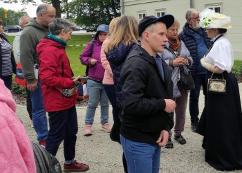Grupa uczestników wycieczki stoi przed budynkiem i słucha przewodniczki ubranej w strój z minionej epoki. Na pierwszym planie widać młodego mężczyznę w czapce, a wokół niego zgromadzonych ludzi ubranych w kolorowe kurtki, rozmawiających i obserwujących otoczenie.