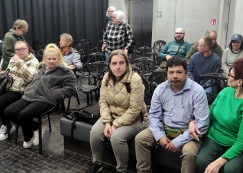 Na zdjęciu znajduje się grupa ludzi siedzących i stojących w sali z nowoczesnym, surowym wystrojem. W tle widać wiele czarnych krzeseł o geometrycznych kształtach ustawionych w rzędach. Ściany są betonowe, a po lewej stronie znajduje się ciemna kotara. Na pierwszym planie siedzi kilka osób — kobieta w jasnej kurtce, mężczyzna w niebieskiej koszuli oraz kobieta w zielonym swetrze. Inni rozmawiają między sobą albo rozsiadają się na miejscach.
