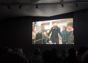 Na tym zdjęciu widzimy ciemną salę kinową lub projekcyjną, w której ludzie siedzą w rzędach i oglądają film wyświetlany na dużym ekranie. Widać tylko sylwetki widzów, ponieważ światło pada głównie z ekranu.  Na ekranie wyświetlona jest scena w jasnym pomieszczeniu, gdzie mężczyzna w historycznym mundurze i hełmie stoi obok starego samochodu, a obok niego stoją dwie młode osoby. U dołu ekranu widać napisy w języku polskim – fragment zdania „który zabierze nas”, co sugeruje, że film ma napisy dla widzów.