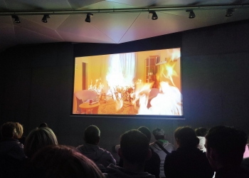 Na tym zdjęciu ponownie widać ciemną salę kinową lub projekcyjną, w której widzowie siedzą i oglądają film wyświetlany na dużym ekranie. Widać jedynie ich sylwetki, co sugeruje przyciemnione oświetlenie, typowe dla oglądania projekcji.  Na ekranie wyświetlona jest scena w jasnym, domowym wnętrzu, w którym meble i część pokoju są objęte płomieniami. Widać m.in. dwa fotele, stolik z kubkiem oraz regał z doniczką. Scena prawdopodobnie przedstawia pożar w mieszkaniu. Kolorystyka ekranu jest intensywna, w ciepłych tonach żółci i pomarańczy, kontrastujących z ciemnym otoczeniem sali.