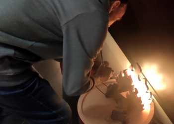 Na zdjęciu widać osobę pochyloną nad jakimś urządzeniem lub instalacją, która emituje światło. Osoba ma na sobie szarą bluzę z kapturem i wydaje się koncentrować na wykonywanej czynności – prawdopodobnie coś podłącza lub reguluje.  Urządzenie lub obiekt w prawym dolnym rogu ma zaokrąglony kształt i jasną powierzchnię, na której widoczne są rozproszone światła – wygląda to jak panel świetlny lub element instalacji artystycznej z wbudowanymi żarówkami lub diodami LED. Część elementów ma faliste lub spiralne kształty. Scena jest oświetlona ciepłym światłem od obiektu, a reszta otoczenia jest ciemniejsza, co skupia uwagę na działaniu osoby i samej instalacji.