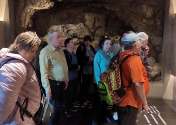 Na zdjęciu widzimy grupę osób stojących w pomieszczeniu, które wygląda jak część muzeum lub wystawy edukacyjnej. Ludzie są ustawieni w kolejce lub skupieni przy ścianie, patrząc na coś znajdującego się po prawej stronie kadru.  W tle widoczna jest skała lub rekonstrukcja naturalnej powierzchni skalnej, na której znajdują się malowidła przedstawiające konie – przypomina to prehistoryczne rysunki naskalne. Oświetlenie jest stonowane, skupione na ścianach, co podkreśla szczegóły rzeźby i malowideł.  Osoby wyglądają na zainteresowane i skupione.