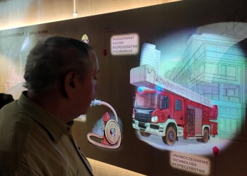 Na zdjęciu widać mężczyznę stojącego blisko ściany ekspozycyjnej w muzeum lub centrum edukacyjnym. Ściana przedstawia kolorową ilustrację poświęconą bezpieczeństwu pożarowemu w miastach. Na grafice widoczny jest czerwony wóz strażacki z wysuwaną drabiną, hydrant z nawiniętym wężem oraz fragmenty miejskich budynków.  Na ścianie są także napisy w języku polskim, m.in.: „ROZBUDOWANY SYSTEM BEZPIECZEŃSTWA POŻAROWEGO” oraz „UNOWOCZEŚNIENIE TECHNOLOGII BEZPIECZEŃSTWA”. Oświetlenie jest skierowane na ekspozycję, podkreślając detale ilustracji i napisy, natomiast reszta sali jest przyciemniona.