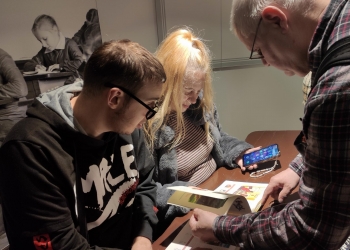 Na zdjęciu widać trzy osoby przy stole podczas zwiedzania muzeum. Młody mężczyzna w okularach i ciemnej bluzie oraz blond kobieta skupiają uwagę na otwartej książce. Obok nich starszy mężczyzna w koszuli w kratę pokazuje coś na jednej ze stron publikacji. Kobieta trzyma w dłoni telefon komórkowy z widocznym kolorowym ekranem.  W tle znajduje się czarno-białe zdjęcie przedstawiające dzieci podczas nauki w klasie, co nadaje miejscu historyczny i edukacyjny charakter. Atmosfera na zdjęciu wygląda na spokojną i pełną zainteresowania wspólnym odkrywaniem informacji.