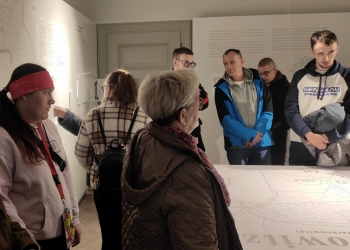Na zdjęciu widać grupę osób zwiedzających nowoczesną wystawę muzealną. Uczestnicy stoją wokół dużego stołu lub planszy z mapą i opisami historycznymi. W pomieszczeniu znajdują się jasne ściany z tekstami informacyjnymi oraz ilustracjami odnoszącymi się do historii miejsca. Po lewej stronie stoi młoda kobieta w jasnej bluzie i różowej opasce, obserwująca ekspozycję. W centrum widoczna jest starsza kobieta w okularach oraz kilka innych osób słuchających lub czytających informacje na ścianach. Po prawej stronie stoi młody mężczyzna w biało-granatowej bluzie z kurtką przewieszoną przez ręce. Atmosfera zdjęcia sugeruje wspólne zwiedzanie i zainteresowanie historią prezentowaną na wystawie.