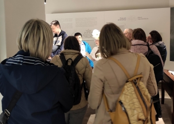 Na zdjęciu widać grupę osób zwiedzających wystawę muzealną poświęconą historii pałacu i dawnej fabryki Giesche. Zwiedzający stoją wokół przewodniczki ubranej w historyczny strój, słuchając jej opowieści i oglądając ekspozycję. Wnętrze jest nowoczesne i jasne, a uczestnicy wycieczki tworzą atmosferę wspólnego zainteresowania historią miejsca.