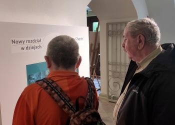 Na zdjęciu widać dwóch mężczyzn oglądających muzealną tablicę informacyjną z napisem „Nowy rozdział w dziejach”. Jeden z nich, ubrany w pomarańczową bluzę i plecak, stoi tyłem do aparatu, podczas gdy starszy mężczyzna obok uważnie przygląda się ekspozycji. Wnętrze muzeum jest jasne i eleganckie, a w tle widoczne są dekoracyjne elementy architektoniczne oraz przejście do kolejnej sali wystawowej.
