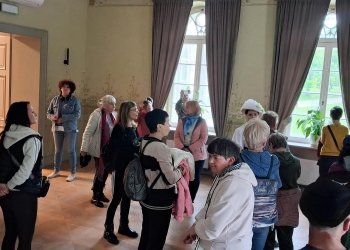 W przestronnej sali muzealnej zgromadziła się grupa zwiedzających, którzy słuchają przewodniczki ubranej w stylizowany historyczny strój z białym kapeluszem. Wnętrze ozdabiają wysokie okna z długimi zasłonami oraz dekoracyjne ściany, co nadaje miejscu elegancki i zabytkowy charakter. Część uczestników rozmawia i obserwuje otoczenie, a przez okna widać zielony park znajdujący się na zewnątrz budynku.