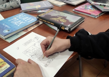 Na zdjęciu widać osobę rysującą komiks na przygotowanej karcie. Na stole leżą liczne komiksy i książki, a uczestnik skupia się na uzupełnianiu kolejnych kadrów o rysunki i tekst. Wśród publikacji można zauważyć zarówno kolorowe albumy, jak i powieści graficzne.