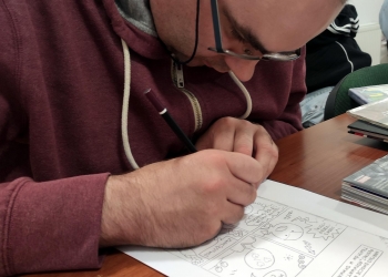 Na zdjęciu widać mężczyznę pochylonego nad kartką papieru, na której tworzy prosty komiks.Skupiony uczestnik warsztatów rysuje ołówkiem kolejne elementy historyjki obrazkowej. Na stole obok leżą komiksy i książki.  