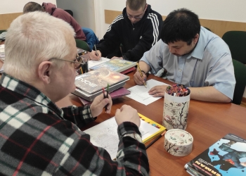 Na zdjęciu kilka osób siedzi przy stole i uczestniczy w kreatywnych warsztatach związanych z komiksem. Uczestnicy skupiają się na tworzeniu własnych ilustracji przy użyciu kredek i ołówków. Na stole znajdują się liczne komiksy, albumy oraz materiały plastyczne. 