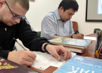 Na zdjęciu dwóch uczestników warsztatów siedzi przy stole i z dużym skupieniem rysuje na przygotowanych kartach pracy związanych z komiksem. Obok nich leżą stosy książek i albumów komiksowych, a także przybory do rysowania. Obaj mężczyźni koncentrują się na tworzeniu własnych ilustracji i uzupełnianiu kadrów komiksowych. 