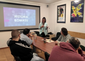 Kobieta stojąca przy ekranie prowadzi prezentację, a na slajdzie widnieje napis „Historia komiksu”. Uczestnicy siedzą przy długim stole i słuchają prowadzącej. Na stole znajdują się książki, komiksy, kubki oraz przybory do rysowania. Na ścianach wiszą kolorowe plakaty artystyczne. 