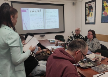 Na zdjęciu grupa uczestników bierze udział w warsztatach poświęconych komiksowi. Prowadząca stoi obok stołu i omawia materiały, a na ekranie wyświetlona jest prezentacja dotycząca emocji przedstawianych za pomocą rysunków i emotikonów. Uczestnicy siedzą przy stole pełnym książek, komiksów i przyborów plastycznych, słuchając prowadzącej lub pracując nad zadaniami.