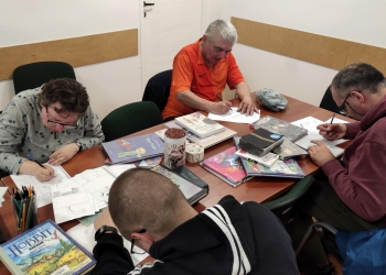 Na zdjęciu kilka osób siedzi przy dużym stole i skupia się na wykonywaniu komiksów. Na stole leżą liczne książki i komiksy, a także pojemniki z kredkami i ołówkami. Uczestnicy pracują w ciszy i z dużym zaangażowaniem, każdy koncentruje się na swoim zadaniu.